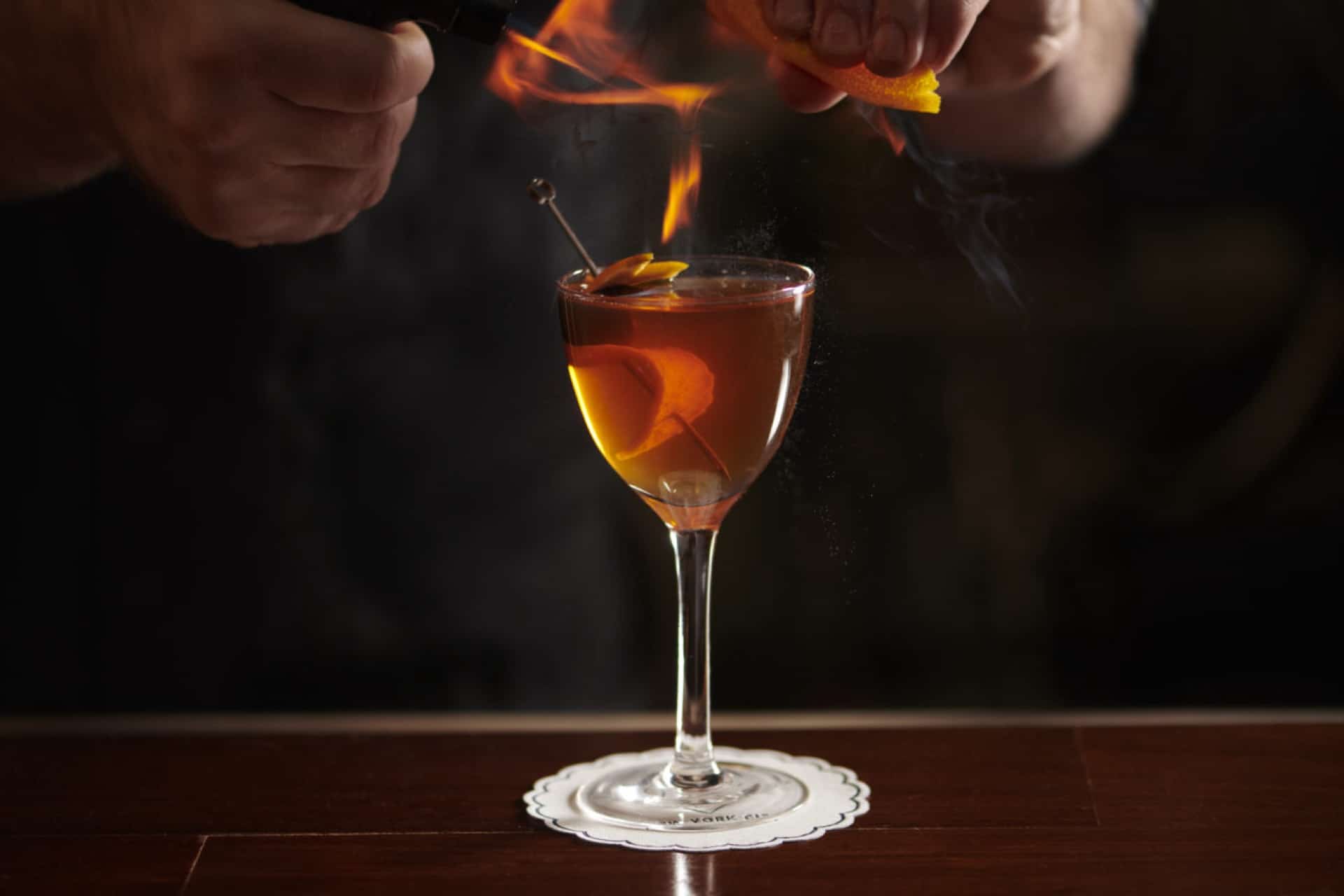 a bartender expressing the oils from an orange over a match above a cocktail